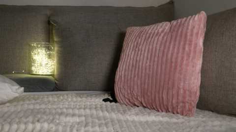 Media: Video of a cozy, modern bedroom corner featuring a plush, ribbed pink pillow on a textured gray upholstered headboard, with a glowing, textured lamp casting a soft yellow light on the white quilted bedspread.