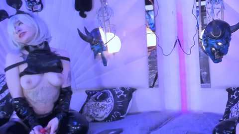 Media: Video of a pale-skinned woman with short, platinum blonde hair, wearing black latex gloves and a black harness, standing in a dimly lit room with purple lighting. The room features dark, gothic decor and hanging skull masks.