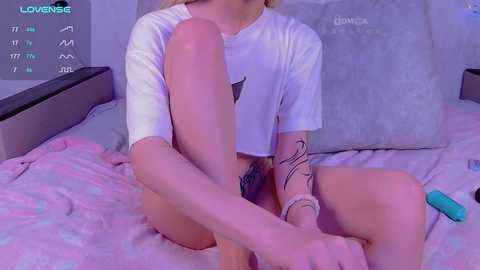 Media: Video of a young woman with fair skin and blonde hair, wearing a white t-shirt and short shorts, sitting on a bed with pink bedding. She has a tattoo on her left forearm.