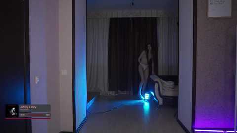 Media: Video of a dimly lit bedroom with a woman in lingerie standing, glowing blue light on the floor.