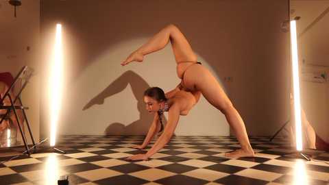 Media: Video of a nude, fit woman performing a challenging yoga pose on a black-and-white checkered floor, surrounded by bright, vertical spotlights casting dramatic shadows against a beige wall.