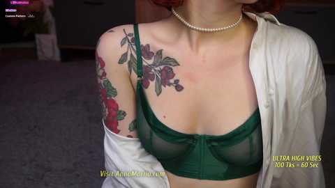 Media: Video of a fair-skinned woman with auburn hair, wearing a sheer green bra, pearl necklace, and open white shirt, showcasing vibrant floral tattoos on her upper arm.