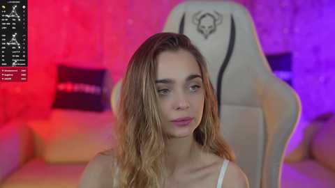 Media: Video of a young woman with light skin and long, wavy brown hair, wearing a white tank top, sitting in a beige gaming chair under vibrant purple and red lighting.