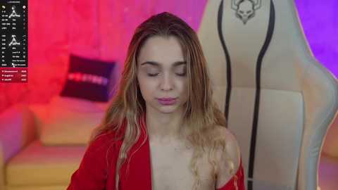 Media: Video of a young woman with long, wavy blonde hair and fair skin, wearing a red robe, sitting in a beige gaming chair, in a dimly lit room with red and purple lighting.