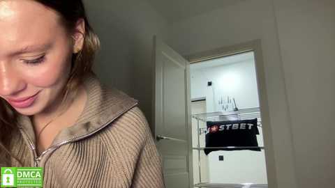 Media: Video of a smiling woman with light skin, wearing a ribbed, light brown cardigan, standing in a dimly lit hallway with white walls. In the background, a laundry room with a drying rack and black clothes is visible.