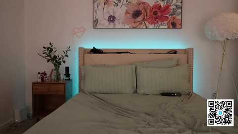 Media: Video of a modern bedroom with a wooden headboard, a beige bedspread, two pillows, a nightstand with a vase, a framed floral painting, and a large, fluffy lamp.