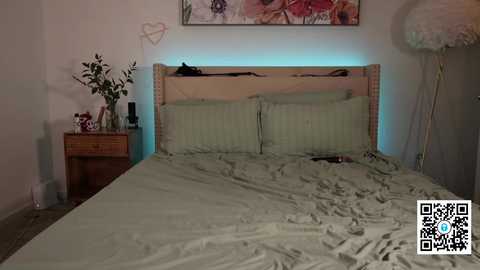 Media: A video of a messy bedroom with a beige bed, green pillows, a wooden nightstand with a vase, and a colorful floral painting on the wall.