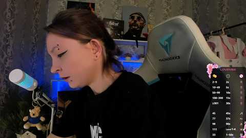 Media: Video of a young woman with pale skin and brown hair in a black shirt, using a blue and white microphone in a dimly lit room with gaming chairs and posters on the wall.