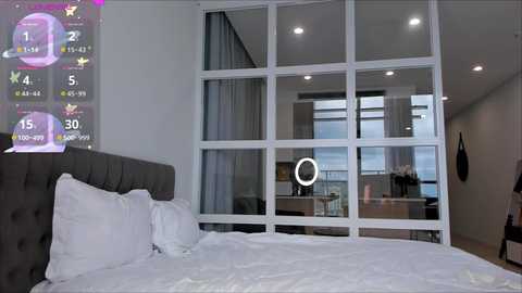 Media: Video of a modern, minimalist bedroom with a gray upholstered headboard, white bed linens, and a large floor-to-ceiling window with a built-in round mirror. Virtual fitness display overlays the left side, showing exercise stats.