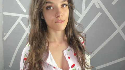 Media: Video of a young woman with long, wavy brown hair, fair skin, and brown eyes, wearing a white shirt with red cherries. She has a small nose piercing. Background shows a geometric pattern in grey and white.