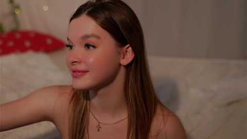 Media: Video of a young Caucasian woman with long, straight brown hair, fair skin, and a slender physique. She wears a gold necklace and smiles gently. Background shows a white bed with a red pillow.