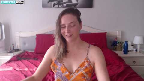 Media: A video of a smiling, fair-skinned woman with brown hair in a floral dress, sitting on a bed with red sheets in a softly lit bedroom.