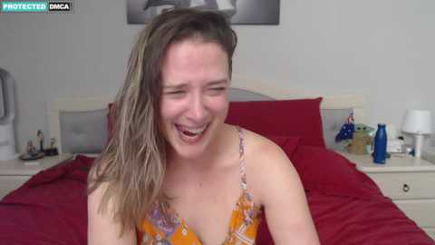 Media: A video shows a smiling, fair-skinned woman with wet, shoulder-length brown hair in a floral dress, sitting on a bed with red sheets and white nightstands in the background.