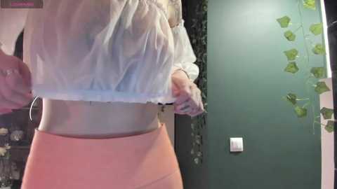 Media: Video of a light-skinned woman lifting her sheer, white blouse to reveal her midriff and peach-colored high-waisted pants. She's indoors with a green wall and hanging ivy.