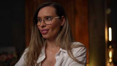 Media: Video of a light-skinned woman with long blonde hair, wearing glasses, a white shirt, and a necklace, in a dimly lit room with wooden panels.