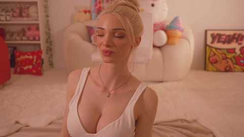 Media: Video of a blonde woman with fair skin, wearing a low-cut white tank top, sitting on a beige carpet in a cozy, colorful room with plush toys and comic book posters.