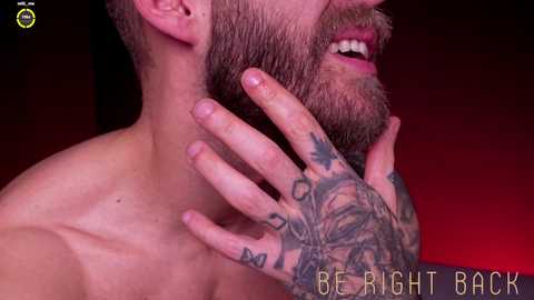 Media: Video of a shirtless man with a muscular build, fair skin, and a full beard. His right hand, adorned with tattoos, touches his face. Background is a gradient of dark red to black. Text reads \"BE RIGHT BACK.\