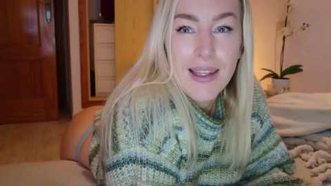 Media: Video of a smiling, fair-skinned blonde woman with straight hair, wearing a green and white striped sweater, in a cozy bedroom with a wooden door, white bed, and orchid.