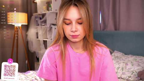 Media: Video of a young Caucasian girl with long, straight blonde hair, wearing a pink t-shirt, sitting on a bed with floral sheets, in a cozy room with a white lamp and purple curtains.