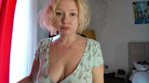 Media: A video of a fair-skinned blonde woman with a short bob haircut, wearing a low-cut floral dress, standing in a bedroom with a bed, red curtains, and a colorful poster on the wall.