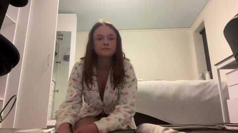 Media: Video of a young woman with long brown hair, wearing a floral-patterned pajama set, sitting on a white bed in a modestly furnished bedroom.