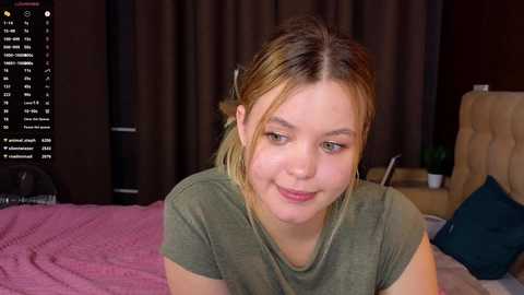 Media: Video of a young woman with light skin, blonde hair in a ponytail, wearing a green shirt, smiling, sitting on a pink bed with a brown headboard, dark curtains in the background.