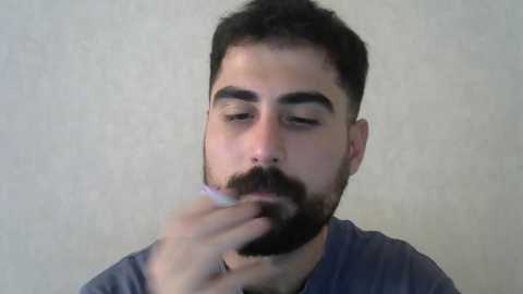 Media: A video of a bearded man with dark hair, wearing a dark blue shirt, inhaling smoke from a white cigarette held in his right hand, against a plain, beige background.