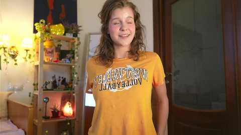 Media: Video of a smiling, curly-haired teenager in a yellow \"Helsinki University\" T-shirt, standing in a cozy, warmly lit room with bookshelves, a fireplace, and a wooden door.