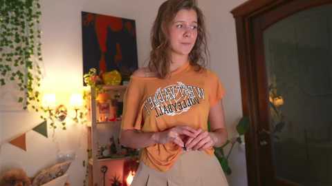 Media: Video of a young woman with messy hair, wearing an orange \"Heavy Metal\" t-shirt and beige pants, standing in a cozy room with a wooden door, green ivy, and warm lighting.