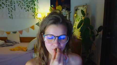 Media: Video of a young woman with curly hair, wearing glasses, and praying, in a dimly lit bedroom with green ivy, a yellow lamp, and a potted plant.