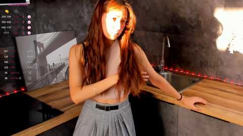 Media: Video of a young woman with long red hair, wearing a grey skirt, standing in a dimly lit kitchen with modern decor, including a wooden countertop, black appliances, and red LED lights.