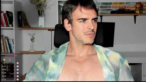 Media: Video of a young man with short dark hair and light skin, wearing a tie-dye shirt, sitting in a home office with bookshelves and a flat-screen TV in the background.