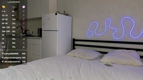 Media: Video of a minimalist bedroom with a white bed, pillows, and a glowing \"buh\" neon sign on the wall. A small kitchen area with a fridge and cabinets is visible in the background.