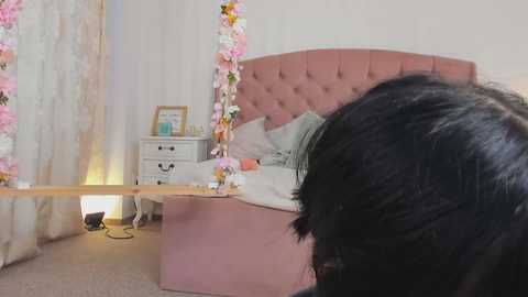 Media: A video shows a young woman with long black hair, partially visible, kneeling near a pink tufted bed adorned with floral decor, surrounded by white walls and curtains.