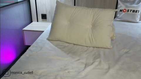 Media: Video of a minimalist bedroom with a large, cream-colored tufted headboard pillow on a white bed, flanked by a white bedside table and a black-and-white abstract wall panel.