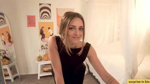 Media: Video of a young woman with light skin, brown hair, and a slim figure, wearing a black sleeveless top. She smiles, leaning forward in a brightly lit, minimalist bedroom with white walls and abstract art.