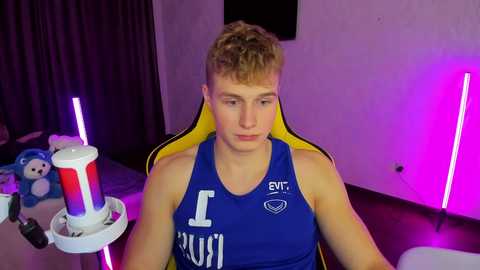 Media: Video of a young, fair-skinned, blonde-haired man in a blue tank top, sitting on a yellow gaming chair, holding a drink. Background features purple lighting, plush toys, and a dark curtain.