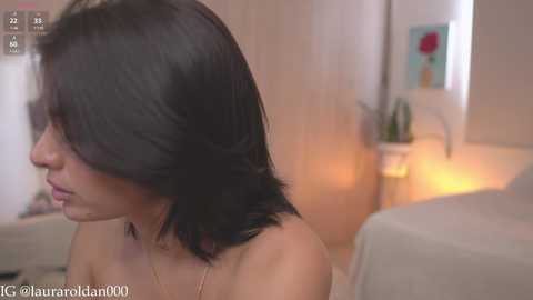 Media: Video of a fair-skinned woman with shoulder-length black hair, partially obscured by a video overlay. She wears a delicate gold necklace and is topless, partially visible in the background.