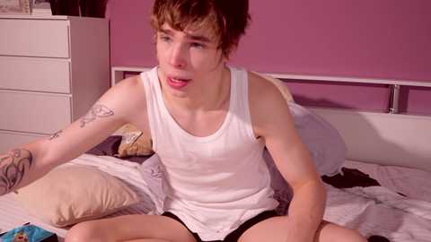 Media: Video of a young man with short brown hair, wearing a white tank top and black shorts, sitting on a bed with messy white bedding, in a purple-painted room.