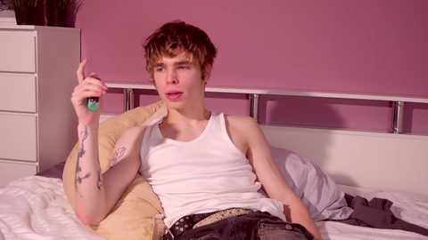 Media: Video of a young, slender, Caucasian male with short brown hair, wearing a white tank top and dark pants, lying on a bed with a pink wall background, holding a green bottle, looking at camera.