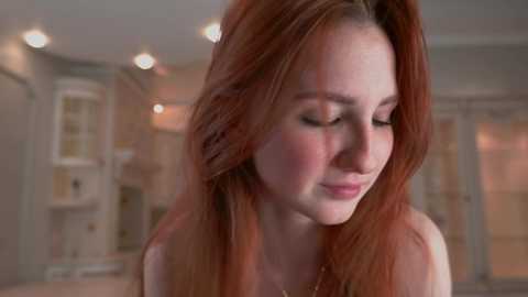 Media: A video of a young woman with fair skin and long, auburn hair, wearing a sleeveless top, standing in a softly lit, softly focused room with beige walls and warm lighting.