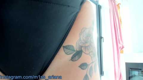 Media: A close-up video of a person's arm, showing a detailed black and gray tattoo of a rose with leaves, partially covered by a black shirt sleeve. The background includes a pink curtain and a window.