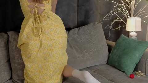 Media: Video of a woman in a yellow, backless dress with a knot detail, standing on a grey couch. Behind her, a green pillow and a white table lamp with a beige shade.