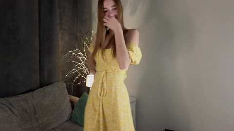 Media: Video of a young woman with straight, light brown hair, wearing a yellow off-shoulder dress, hiding her face with her hands, standing in a dimly lit room with grey walls and a beige sofa.