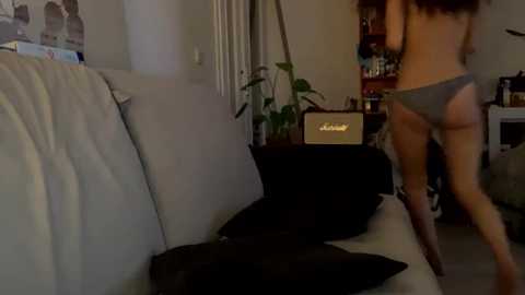 Media: Video of a woman in gray panties, pulling down her black top, in a dimly lit living room with a white couch, black throw pillows, and a vintage amplifier.