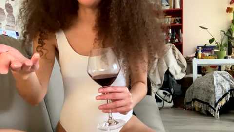 Media: Video of a woman with voluminous curly hair, wearing a white tank top, holding a glass of red wine, in a cozy living room with bookshelves, a grey couch, and a white chair.