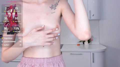 Media: A video of a topless light-skinned person with small breasts, wearing pink shorts, holding their arm, with a tattoo on their chest, in a white kitchen setting.