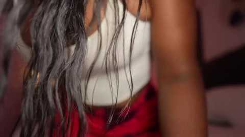 Media: Video of a Black woman with wavy, shoulder-length black hair in a white tank top, wearing a red skirt, against a blurred, muted background.