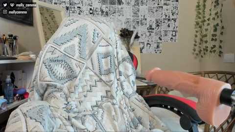 Media: A video shows a person wearing a patterned blanket with geometric designs, being pushed in a wheelchair by another person. The background features a cluttered room with a wall covered in manga posters.