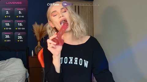 Media: Video of a blonde woman with wavy hair, wearing a black sweatshirt, licking a large, pink dildo in a dimly lit bedroom.